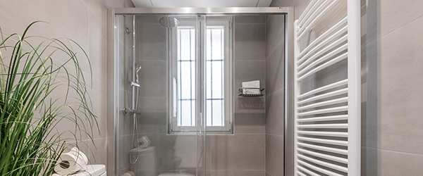 Geflieste Toilette mit großen grauen Fliesen, Pflanze auf einem Holzschemel und Duschkabine mit Glasabtrennung und elektrischem Heizkörper, weißer Handtuchhalter aus Aluminium